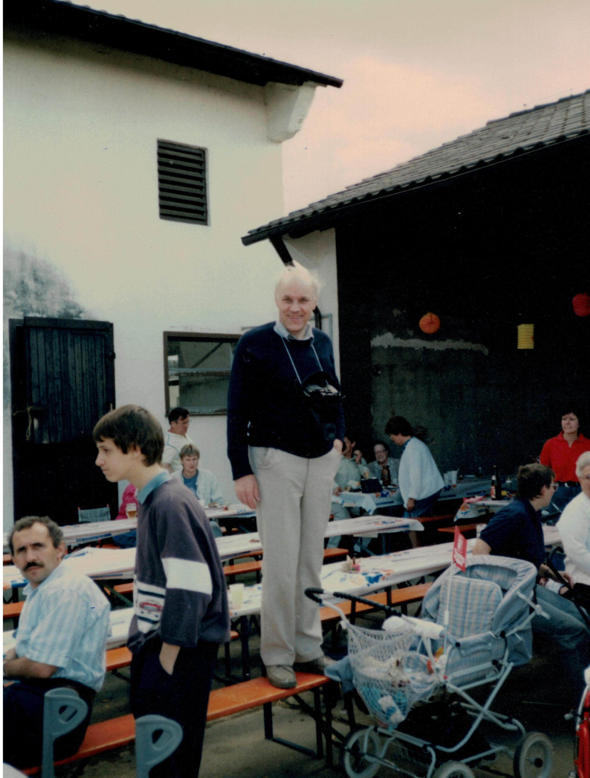 Sommerfest 1988, Dr. Ermert hat eine Kamera umhängen, lächelt und steht dabei auf einer Bierbank in einem Hof, um ihn verteilt Mitglieder, vor ihm ein leere Kinderwagen.