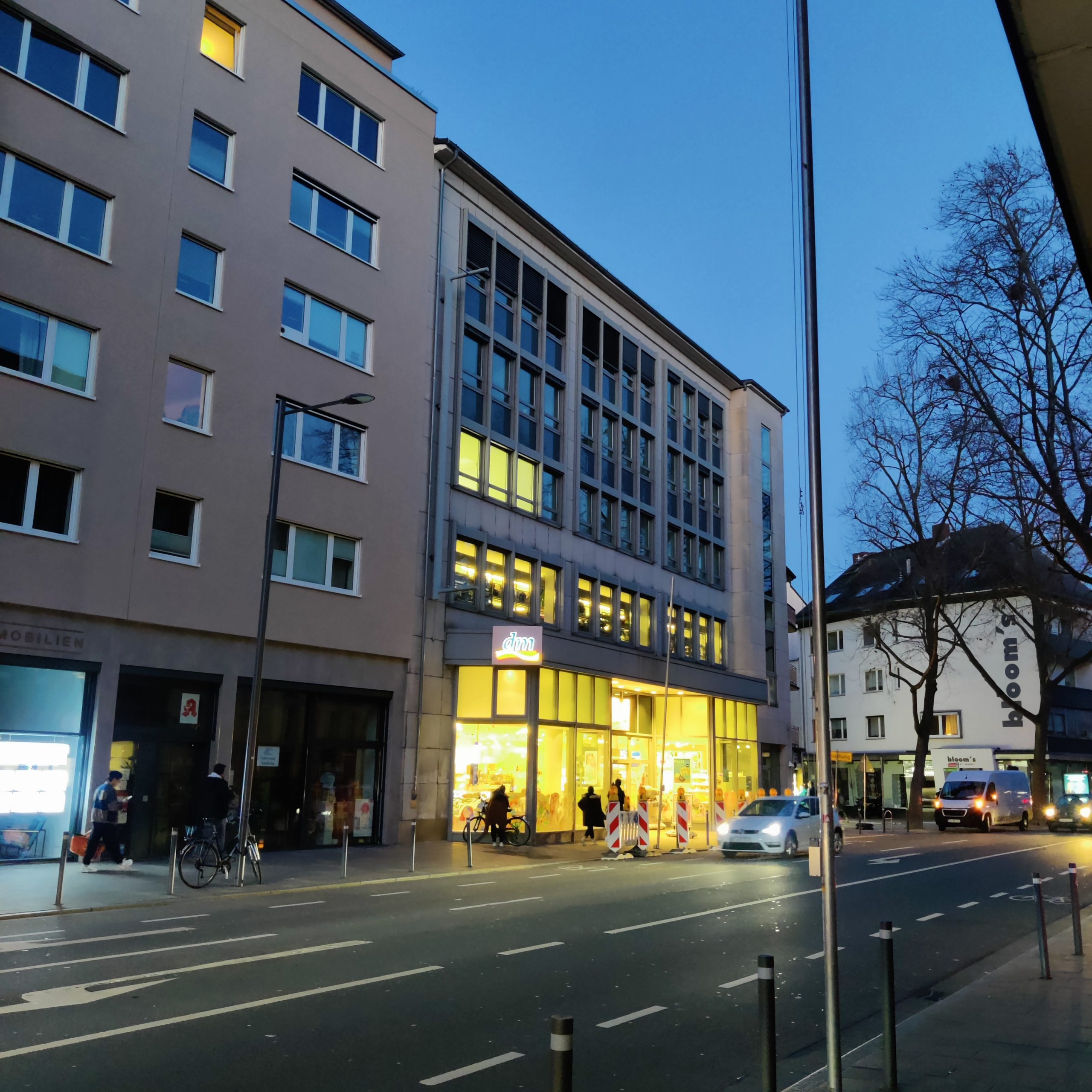 Ein Bürogebäude mit einem DM-Laden im Erdgeschoss in der Dämmerung von der gegenüberliegenden Straßenseite aus fotografiert. Ein Fenster im zweiten Stock ist beleuchtet.